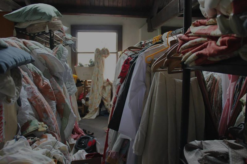 Picture of a room full of textiles either hanging on a rail (on the right) or placed clumsily (on the left), with light from an open window (centre).